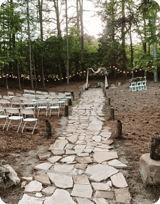 A stone pathway, surrounded by folding chairs, leads to a pergola in the woods at The Glen Wedding Venue at Fablerealm.